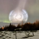 A massive spiral of white light in the night sky with auroras in the background