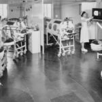 Black and white image of a group of eight patients who are in iron lungs that are arranged in an almost semi-circle manner around a television in a hospital. There are two nurses in the image, one is attending a patient towards the back of the image and another is standing by the television.