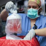 a woman in blue scrubs, gloves and a face mask removed a bagged organ in a jar in preparation for surgery
