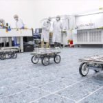 three small rovers with four wheels each pictured in a lab with scientists standing in the background to observe