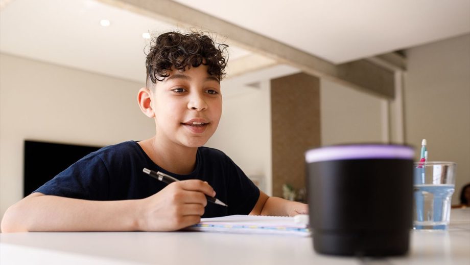 Child with curly brown hair sits at a table with a pencil and looks at a smart speaker on the table, as if to ask it a question