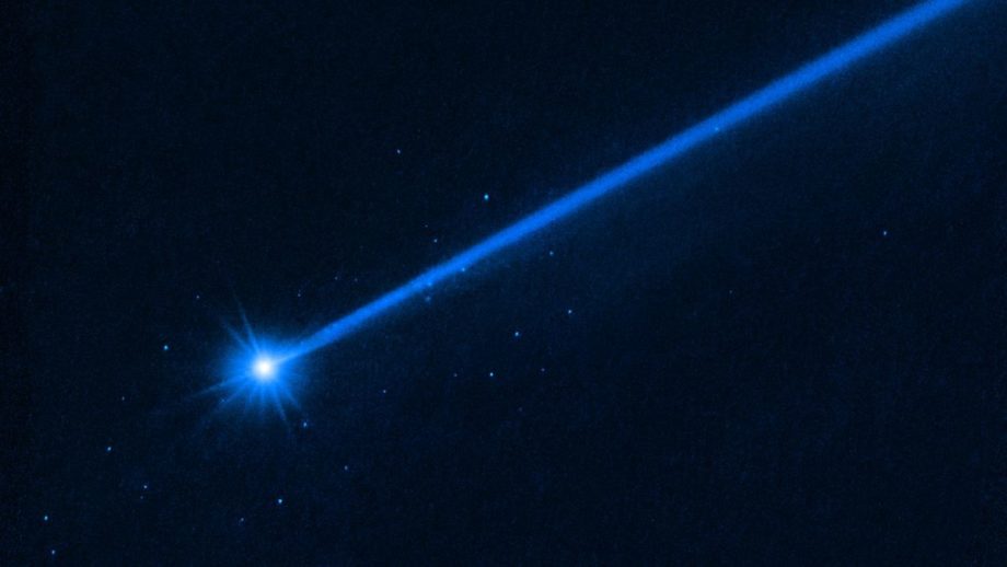 A Hubble telescope image of a bright blue asteroid, trailed by a long blue tail to the right. Small blue dots show boulders blasted away by NASA