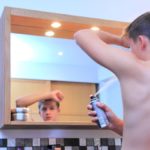 young teen boy using a spray deoderant on his left armpit while he looks in a bathroom mirror