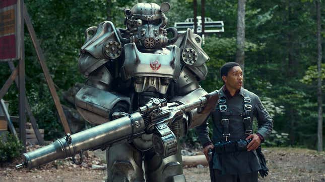 На изображении изображен солдат Fallout в силовой броне, стоящий рядом с человеком в лесу. 