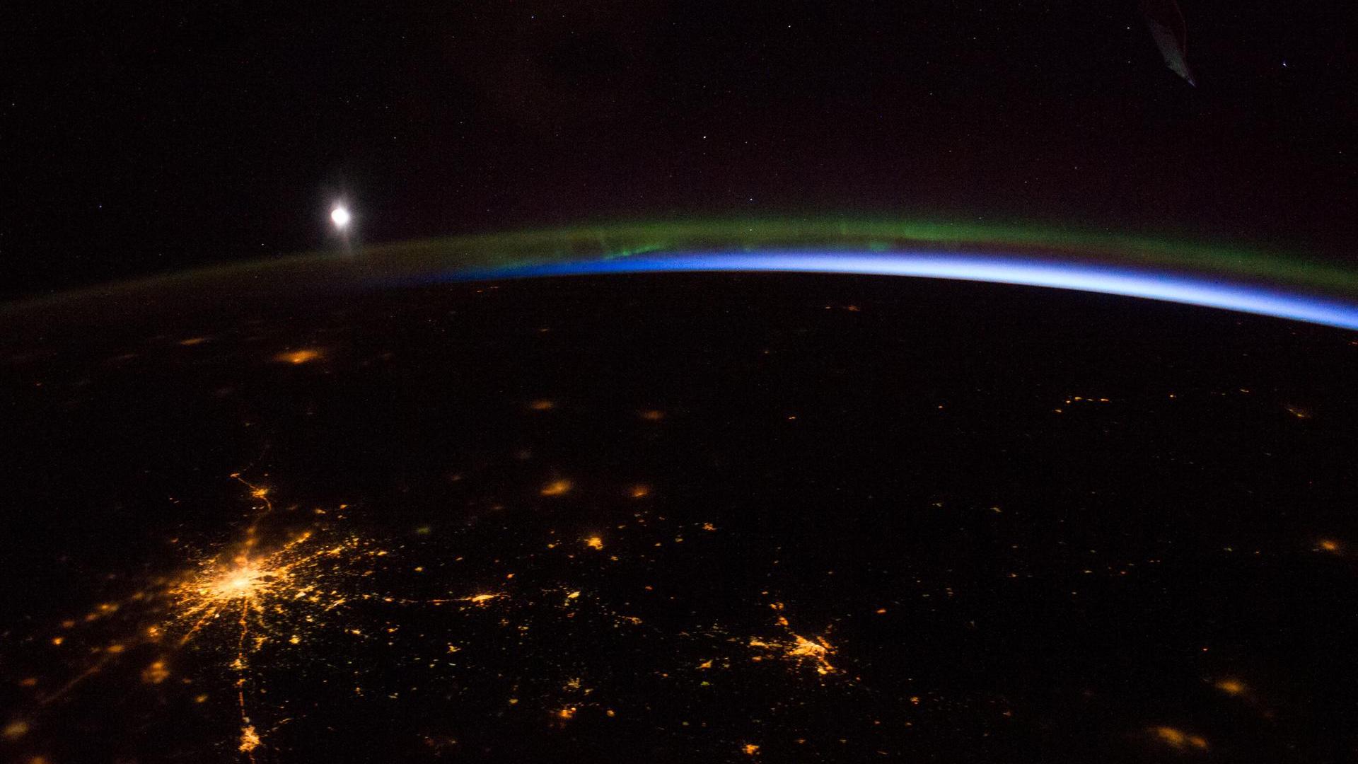 ночная фотография из космоса, показывающая сине-зеленое северное сияние и луну над огнями Москвы.