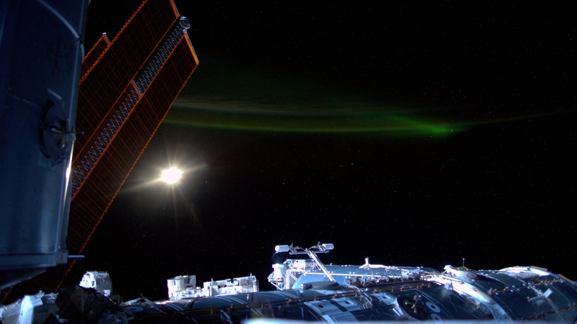 Фотография международной космической станции с Луной и слабым зеленым полярным сиянием на заднем плане.