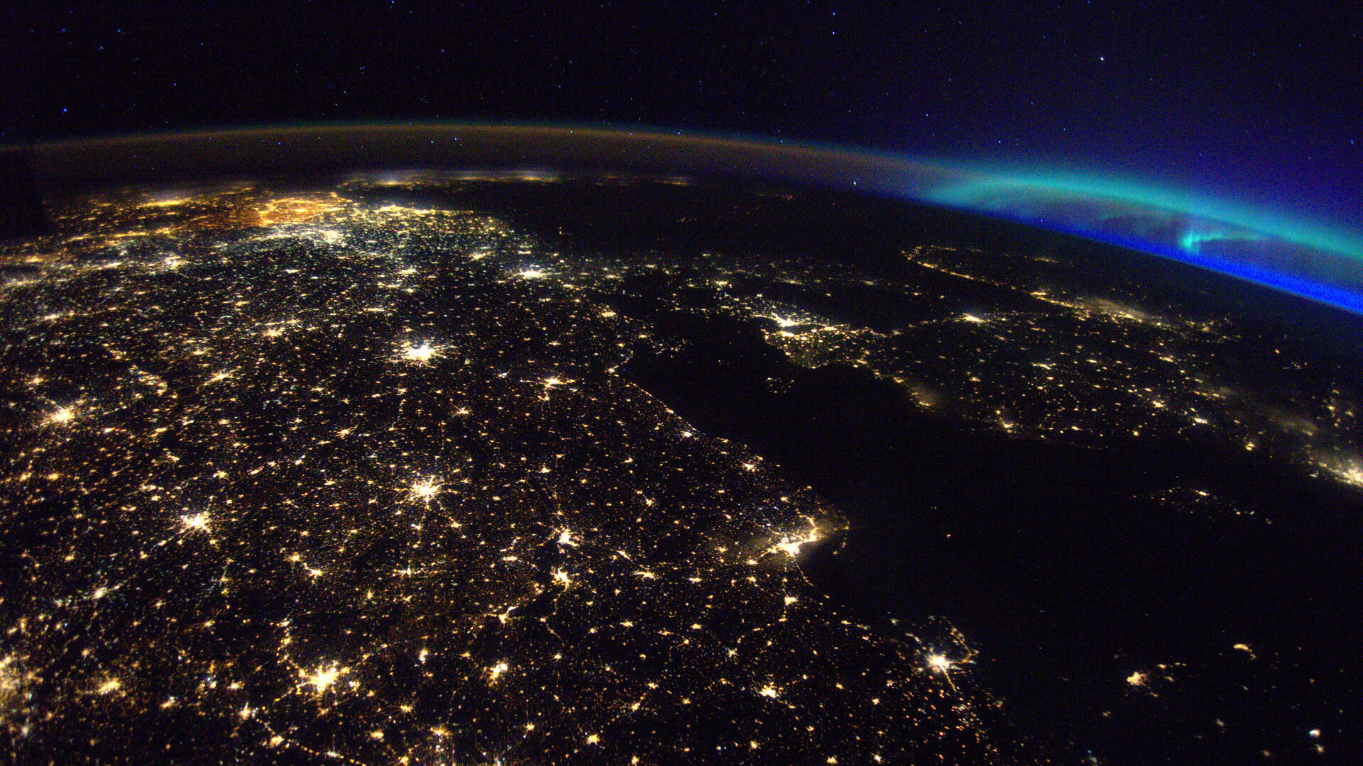 Фотография синего полярного сияния вдалеке с огнями города на переднем плане.