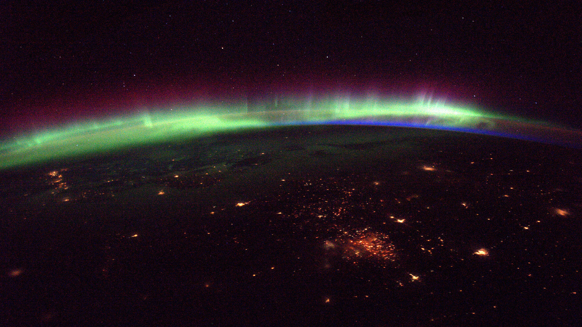 Фотография зеленого, синего и фиолетового сияния над горизонтом земли с красноватыми огнями города на переднем плане.