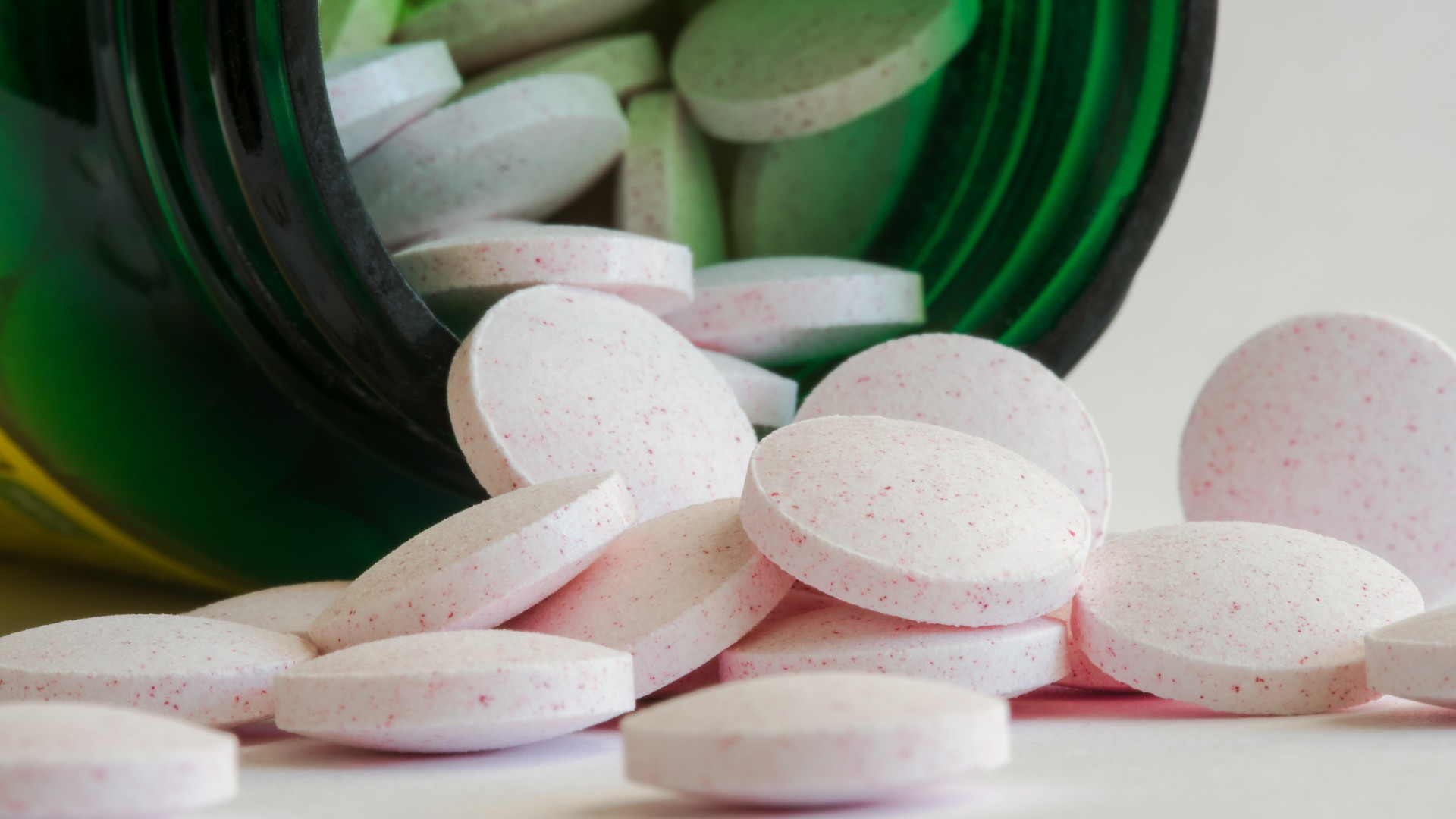 Таблетки мелатонина высыпаются из зеленой бутылки.  Таблетки белого цвета с мелкими розовыми крапинками.  Они похожи на маленькие диски.