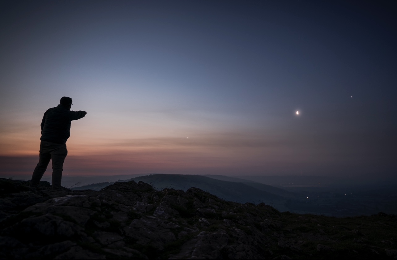 Мужчина стоит на скале и смотрит на расположение шести планет в предрассветном небе.