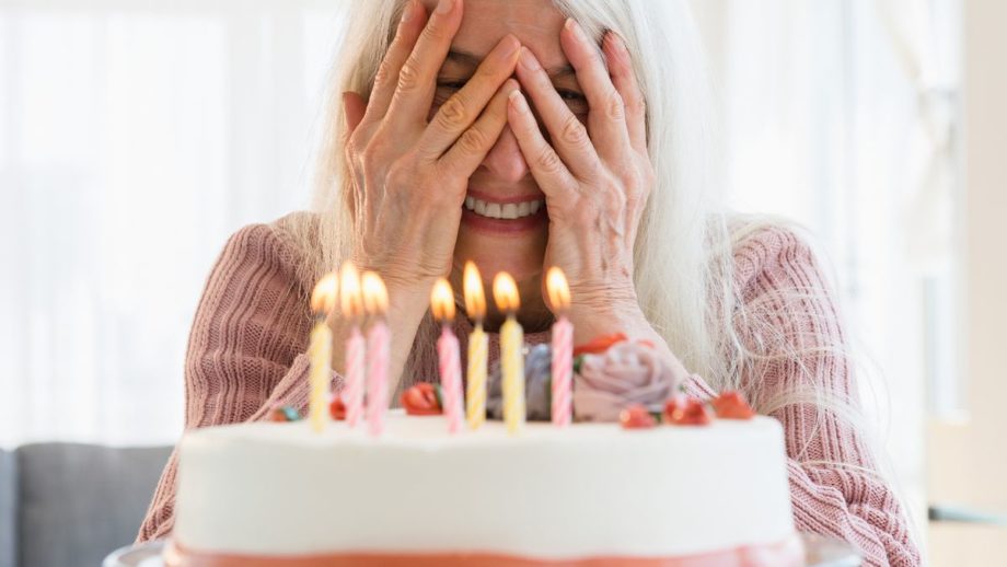 A senior woman with long white hair peeks between her fingers at a birthday cake with candles